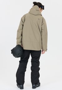 Beige waterdichte jas met een capuchon, zwarte broek, houdt een zwarte helm vast, matte afwerking, losse fit en eenvoudig ontwerp.