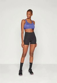 Lila Sport-BH mit Logo und schwarze hoch geschnittene Shorts, kombiniert mit schwarzen Socken und Schuhen mit rosa Akzenten.