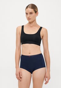 Woman wearing black sports bra and navy high-waist underwear, standing against plain light background with neutral expression.