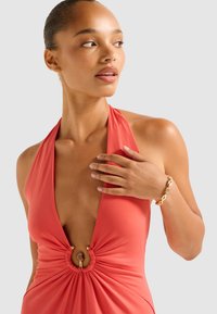 Coral halter-neck swimsuit with a deep V-neck and gathered front featuring a gold circular detail. Model wears a gold link bracelet.