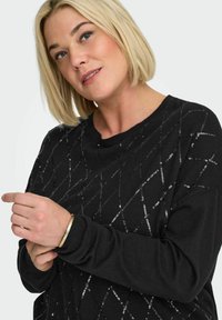 Črni pulover z vzorcem diamantov, ustvarjenim s srebrnimi svinčniki. Ima okrogel izrez in dolge rokave z rebrastimi manšetami.