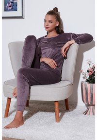 Velveta loungewear set u prigušenoj ljubičastoj boji, s dugim rukavima i uskim hlačama, prikazan na svijetloj stolici uz bijeli zid.