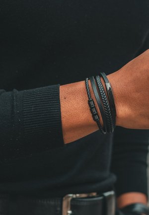 Black leather bracelets in varied textures: braided, smooth, and detailed accents. Worn on a wrist with a black sweater in the background.