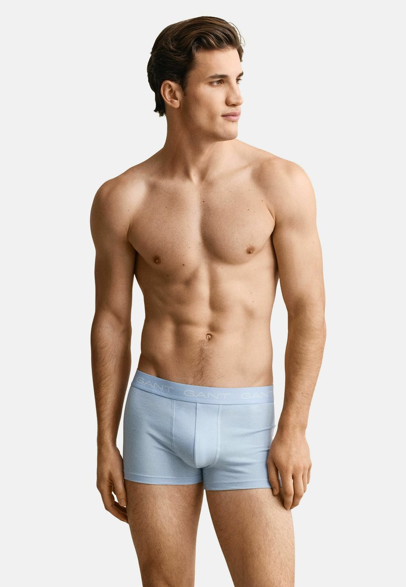 Modèle masculin musclé portant un boxer bleu clair avec une ceinture à logo GANT, debout et regardant vers la droite sur un fond uni.