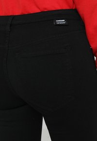 Vaqueros de mezclilla negra con un corte ajustado, que presentan un bolsillo trasero y una etiqueta con la inscripción "DROENIM". La textura parece suave y resistente.