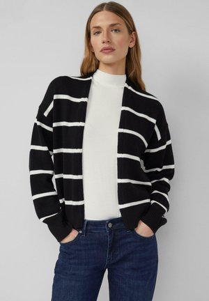 Cardigan nero con righe orizzontali bianche, caratterizzato da una vestibilità ampia, polsini a costine e apertura frontale, indossato sopra un dolcevita bianco.