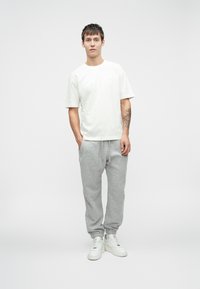 Bijela pamučna majica s kratkim rukavima, u kombinaciji sa sivim trenirkama i bijelim tenisicama. Jednostavan dizajn s opuštenim krojem i elastičnom gumom.