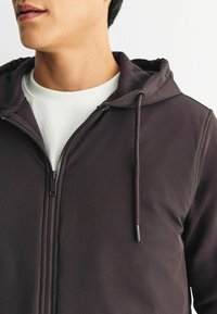 Tumepruun lukuga kapuutsiga jakk pehme tekstuuriga, millel on kapuuts, reguleeritavad tõmblukud ja kontrastsest valgest sisekiht.