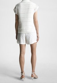 Persona in piedi di spalle che indossa un gilet di maglia bianco senza maniche, pantaloni corti bianchi a fantasia e sandali con tacco bianchi su uno sfondo semplice.