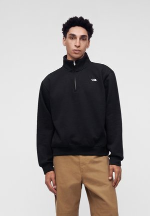 Jeune homme aux cheveux courts et bouclés portant un pull noir à demi-fermeture éclair et un pantalon beige, debout devant un fond clair uni.