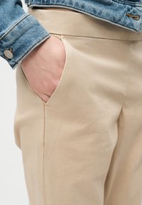 Main dans la poche d'un pantalon beige, portant une veste en denim bleu, montrant la texture du tissu et les détails de couture sur les vêtements.