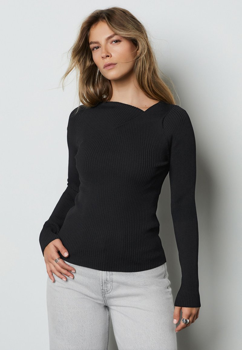 Femme aux longs cheveux blonds portant un haut noir côtelé moulant à manches longues et un jean gris clair, debout devant un fond clair uni.
