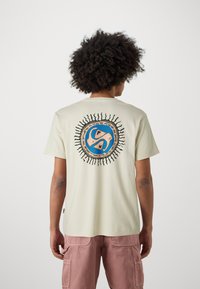 Cream-colored cotton t-shirt with short sleeves features a circular blue graphic on the back, outlined in black with sun-ray accents.