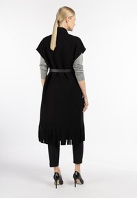 Zwarte wollen vest met een tailleband, korte mouwen en een franjerand, gecombineerd met een grijze langemouwen top en zwarte broek met hakken.