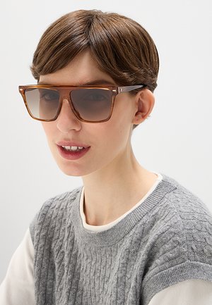 Donna con capelli castani corti che indossa grandi occhiali da sole marroni con lenti sfumate e un maglione grigio lavorato a maglia senza maniche sopra una maglia bianca a maniche lunghe.