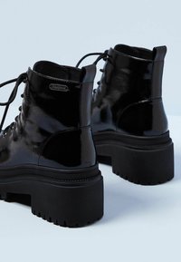 Bottes à lacets en cuir verni noir avec semelles épaisses à plateforme et languettes, vues de dos sur un fond clair.