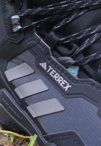 Crne planinarske cipele s mrežastim gornjim dijelom, tri sive pruge, ojačani nos i istaknuti "TERREX" logo u bijeloj boji. Izdržljiv dizajn.