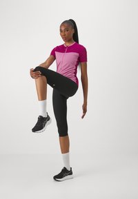 Purpurinės ir rožinės spalvų sportinė palaidinė su pusiau užtrauktuku, juodos kapri kelnės ir juodi sportbačiai. Matomos baltos kulkšnies kojinės. Lieknas siluetas.