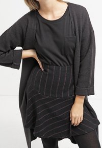 Long cardigan noir sur un t-shirt noir avec une poche ; associé à une jupe noire avec des rayures rouges horizontales et un tissu texturé.
