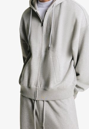 Mand iført lysegrå hættetrøje med lynlås og matchende joggingbukser med hvid t-shirt under, stående mod hvid baggrund.
