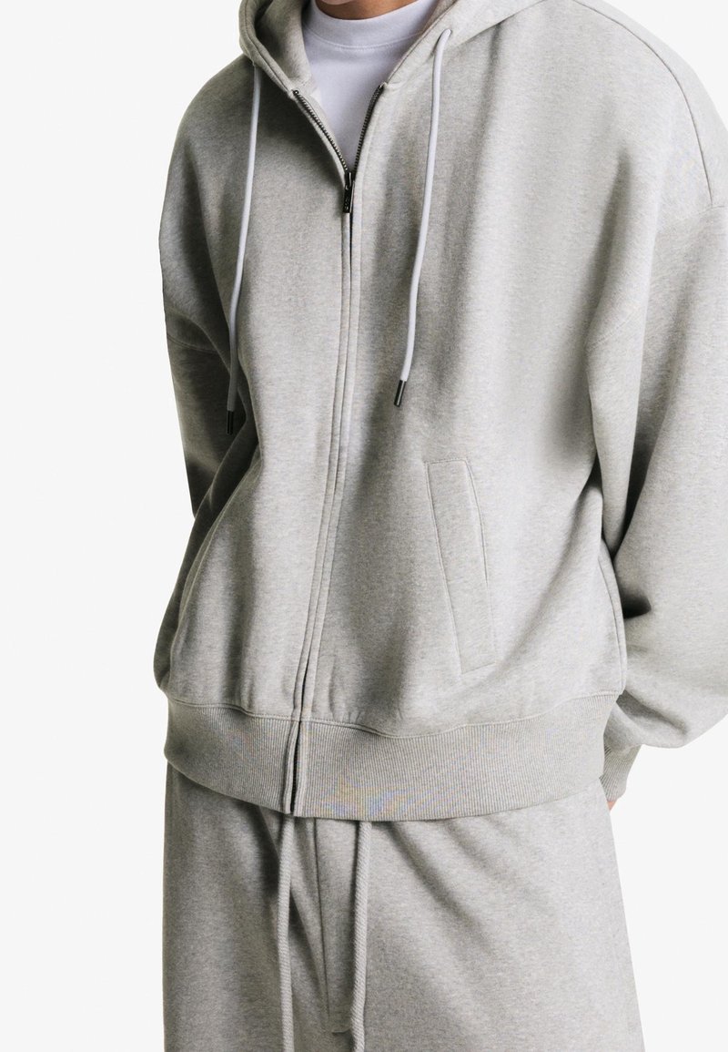 Homme portant un sweat à capuche zippé gris clair et un pantalon de survêtement assorti avec un t-shirt blanc en dessous, debout devant un fond blanc.
