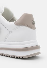Talons de baskets blanches avec accent en cuir beige et logo de marque rond, présentant une semelle épaisse avec des couches beige et blanche et une semelle extérieure striée.
