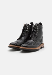 Botas de cuero negro con diseño tipo wingtip, intrincadas perforaciones y suela de madera marrón. Presentan cordones en contraste y una pestaña en el talón.