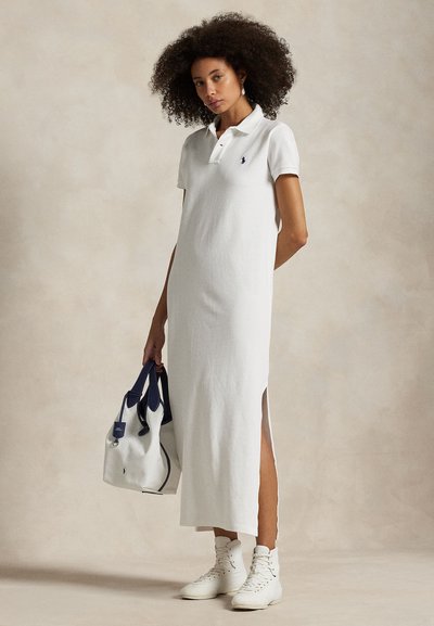 Valkoinen polopuku, jossa suora leikkaus, lyhyet hihat, sivuhalkiot ja vaakuna. Yhdistetty valkoiseen käsilaukkuun ja valkoisiin korkeavartisiin tennareihin.