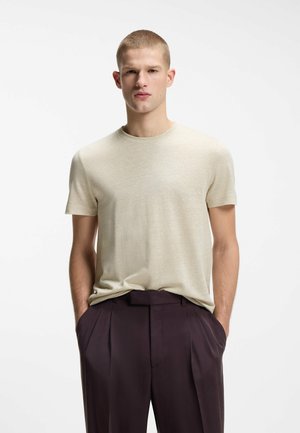 Junger Mann steht mit den Händen in den Taschen, trägt ein beiges kurzärmeliges T-Shirt und dunkle plissierte Hosen vor weißem Hintergrund.