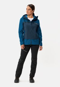 Blue and navy waterproof jacket with a hood, front zipper, and two side pockets, paired with black pants featuring textured knee accents.