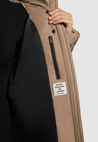 Hånd der holder en åben beige jakke, der viser sort indvendig foret, lynlåslomme og et mærke med teksten "Krammet er lavet af stof".