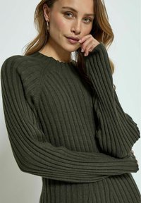 Pull en mailles côtelées de couleur vert foncé en tissu doux, doté d'un col rond et de manches allongées qui créent un aspect texturé et ajusté.