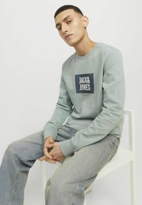 Sudadera verde menta con cuello redondo, que presenta un gráfico cuadrado con "JACK & JONES" en texto blanco. Vaqueros azules, sentado en una silla blanca.