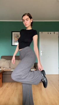 Top preto ajustado, calças cinza com efeito canelado e botins pretos com fivelas prateadas. Ambiente interior com parede verde e sofá bege visíveis.