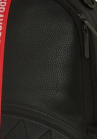 Zaino nero con texture, dotato di una tracolla rossa, chiusura con zip e motivo geometrico sul fronte. Materiale resistente e design elegante.