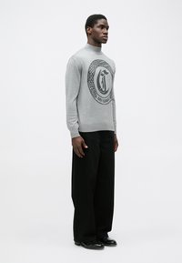 Pulover gri cu un design de logo rotund ce are textul "JUST CAVALLI". Modelul îl asortează cu pantaloni largi negri și pantofi din piele.