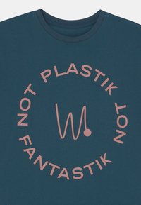 T-shirt teal feita de algodão com mangas curtas. Apresenta um design circular em texto rosa com a inscrição "NOT PLASTIK" e um "W" estilizado.