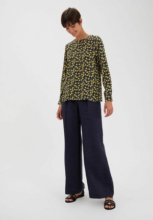 Haut noir à manches longues avec des motifs de pois jaunes, associé à un pantalon navy ample. Le mannequin se tient dans une posture détendue, portant des sandales noires.