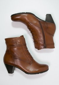 Botas de cuero marrón hasta el tobillo con punta redonda, parte delantera con cordones y cremallera lateral. Presenta un tacón apilado y una textura suave.
