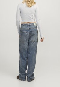 Frau in lockeren blauen Jeans und einem hellgrauen langärmeligen Crop-Top, die mit dem Rücken zu einer schlichten weißen Wand steht.