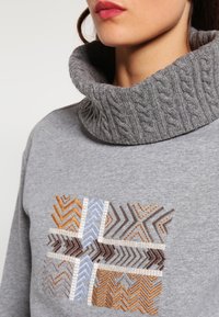 Grijze trui met een dikke, gestructureerde col. Heeft een kleurrijk geometrisch patroon in bruin, blauw en oranje aan de voorkant.