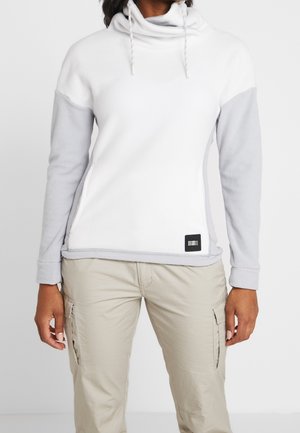 Person iført hvid pullover med lysegrå ærmer og beige cargobukser, står mod en ensfarvet baggrund.