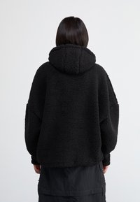 Hoodie en polaire noire avec une surface douce et texturée, coupe surdimensionnée et grande capuche. Équipé d'épaules tombantes et d'un ourlet allongé.