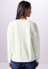 Lichtgrijze sweatshirt met lange mouwen, ribbelachtige halslijn en verlaagde schouders, met een centrale naad op de rug, gedragen met blauwe jeans.