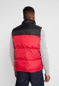 Rote und schwarze Steppweste mit hohem Kragen, geprägter Textur und leichtem Material, getragen über einem grauen Pullover. Rückansicht gezeigt.