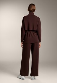 Barna, cropped pulóver magas gallérral és elasztikus derékkal, laza szabású barna nadrággal párosítva. Puha anyag sima textúrával.