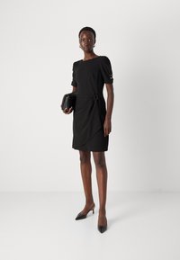 Abito nero con maniche a palloncino corte, dettaglio annodato in vita e pannello drappeggiato sul davanti. Il modello tiene una pochette nera; abbinato a tacchi a punta.
