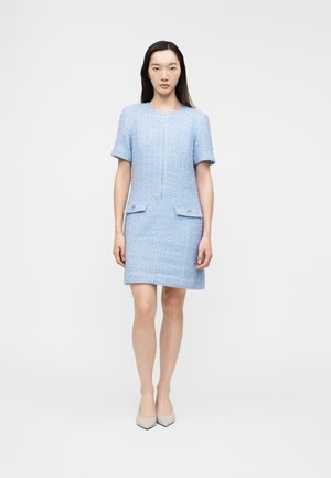 Woman wearing a light blue textured short-sleeve dress with front pockets and a concealed front zipper, paired with beige pointed shoes.