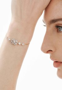Pulsera de oro con eslabones cuadrados y en forma de diamante, con acentos de piedras preciosas claras, llevada en una muñeca. Primer plano de la pulsera y la mano.