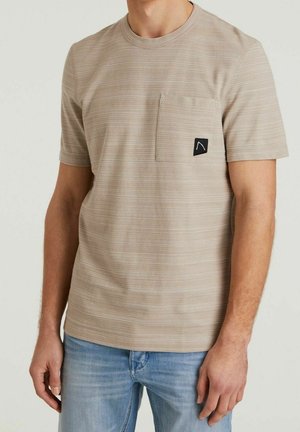 Hombre con camiseta beige de manga corta acanalada con bolsillo en el pecho y parche negro con logotipo, combinado con jeans de mezclilla azul claro.